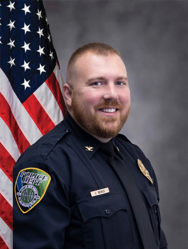 Male police officer in dark uniform with badge and name tag, smiling beside a U.S. flag.